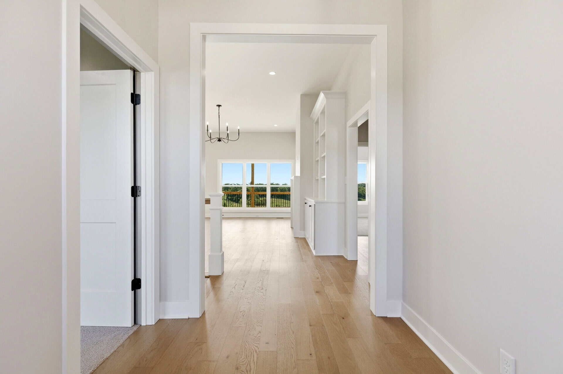 Bright hallway with light wood flooring, white walls and door frames; open doors lead to a sunlit room with a chandelier and large windows showing greenery.