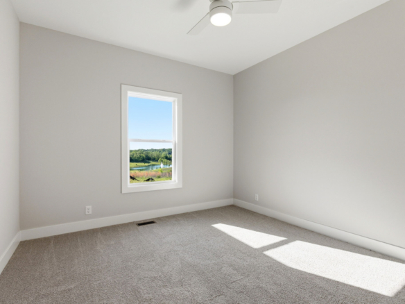 Empty sunlit bedroom with light gray walls, carpeted floor, and a window showing trees outside; ceiling fan overhead.