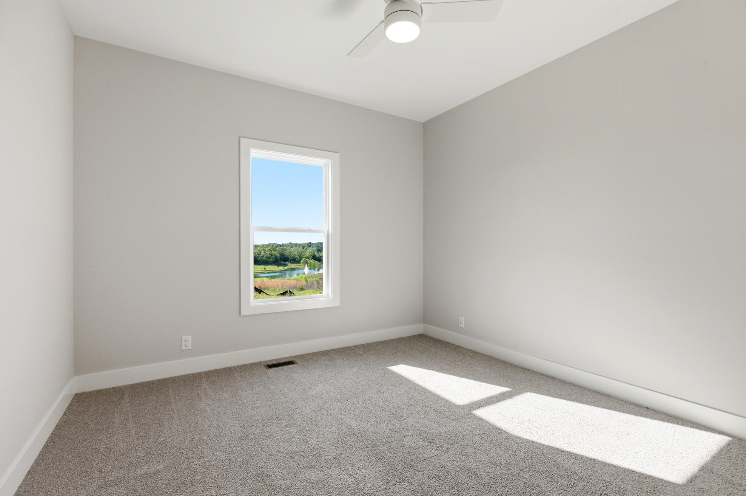 Empty sunlit bedroom with light gray walls, carpeted floor, and a window showing trees outside; ceiling fan overhead.
