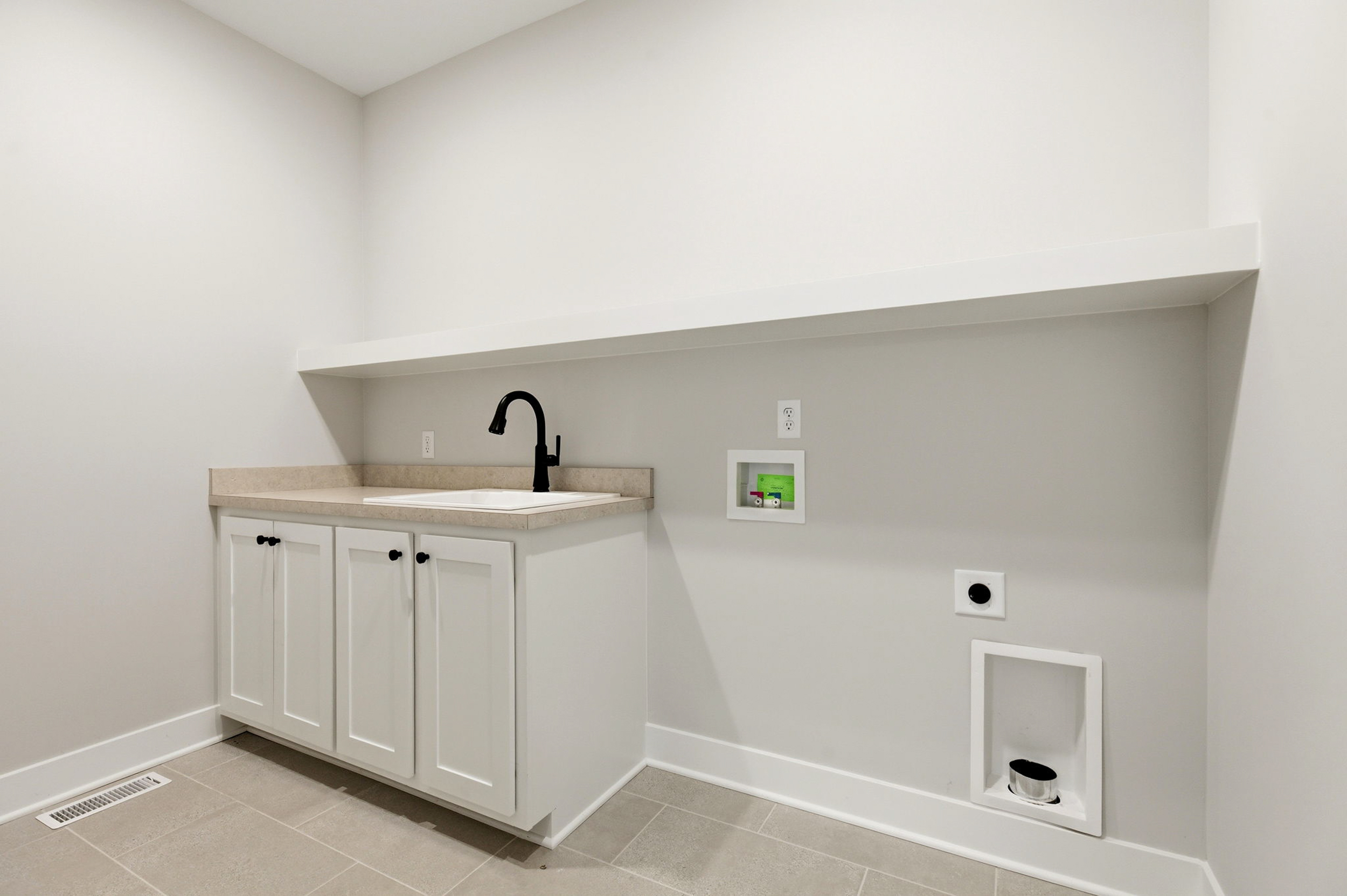 Modern white kitchen/utility alcove with beige countertop, black gooseneck faucet, and white cabinet base under a long shelf.