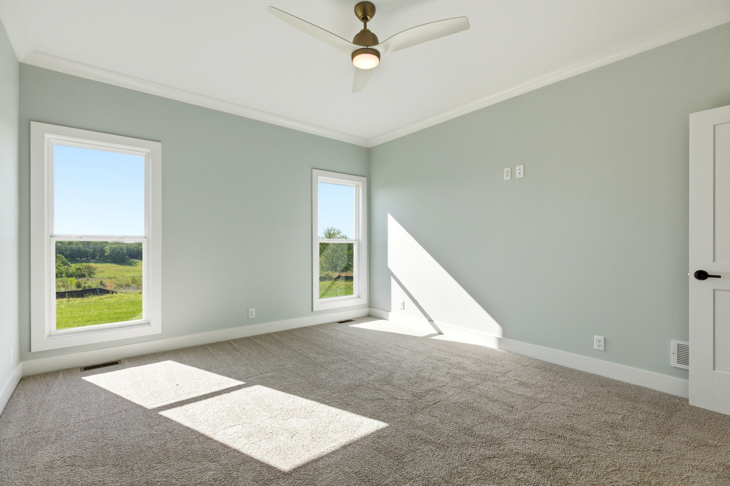 Empty sunlit room with light gray walls, white trim, and beige carpet; two large windows show a green landscape outside, and a modern ceiling fan hangs from the white ceiling.