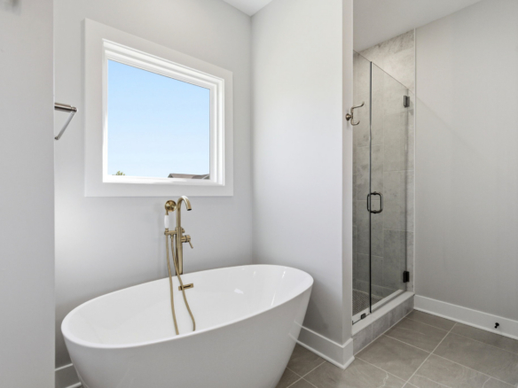 Bright bathroom with a white freestanding tub and brass faucet, window above, and a glass-enclosed shower to the right.