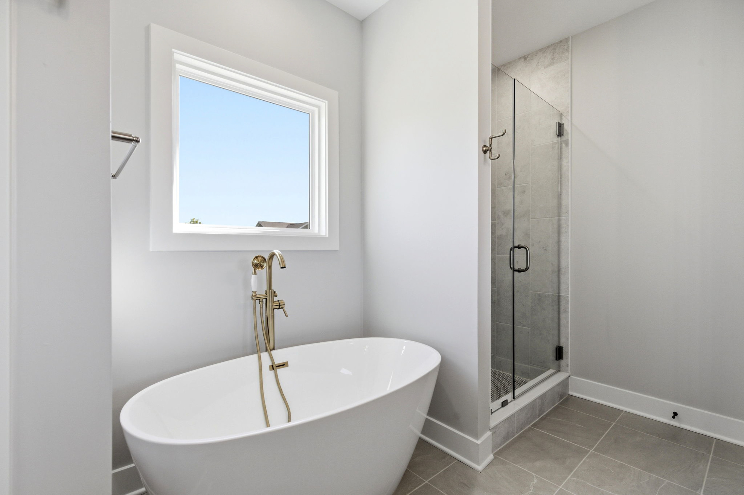 Bright bathroom with a white freestanding tub and brass faucet, window above, and a glass-enclosed shower to the right.