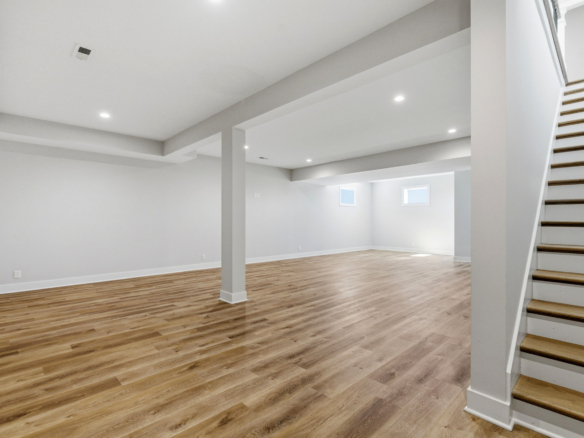 Open empty basement with light wood flooring, white walls, and a right-side wooden staircase.