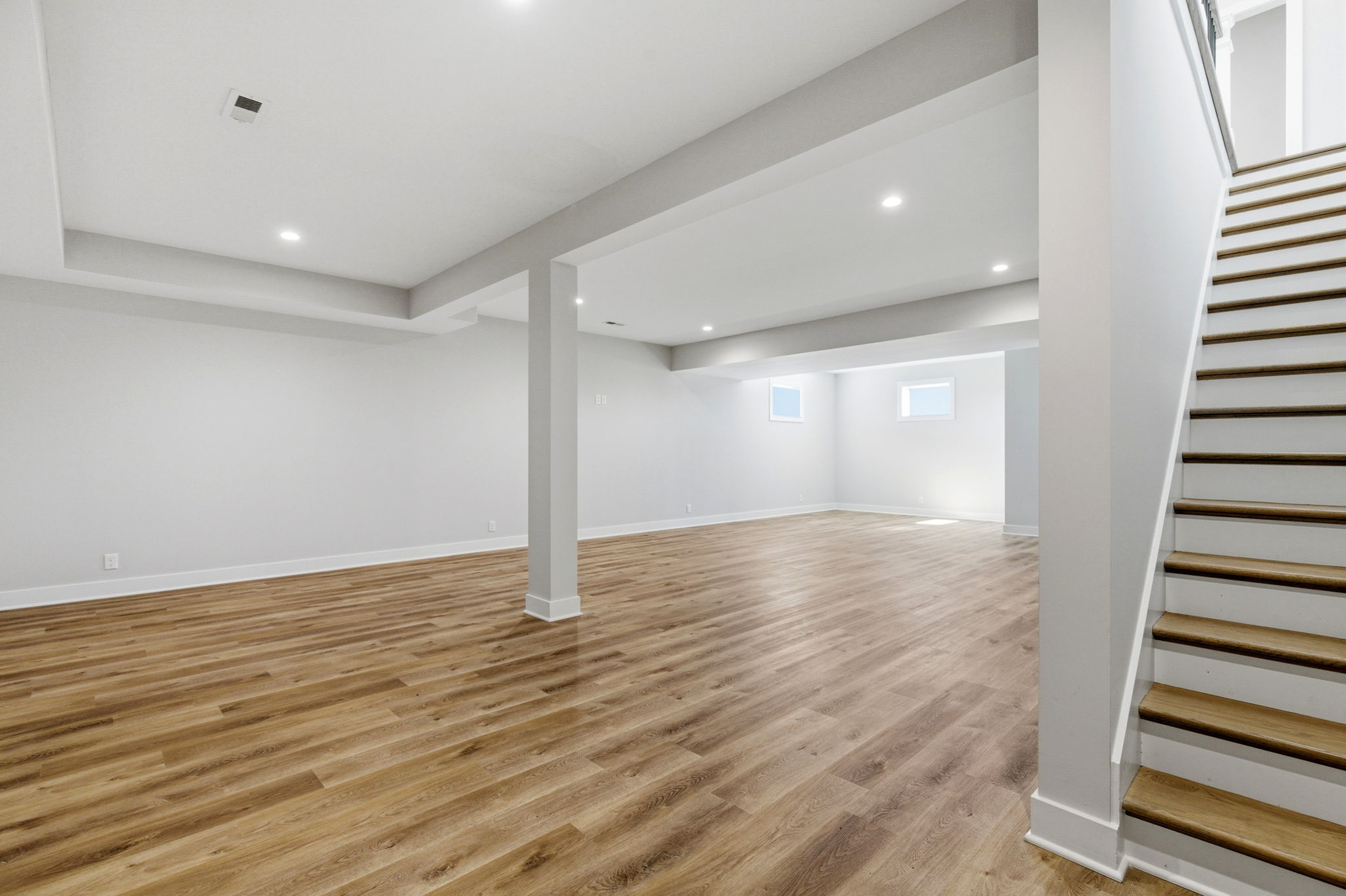 Open empty basement with light wood flooring, white walls, and a right-side wooden staircase.