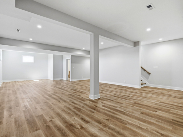 Open, empty basement with light gray walls, wood-look flooring, exposed columns, and recessed ceiling lights.
