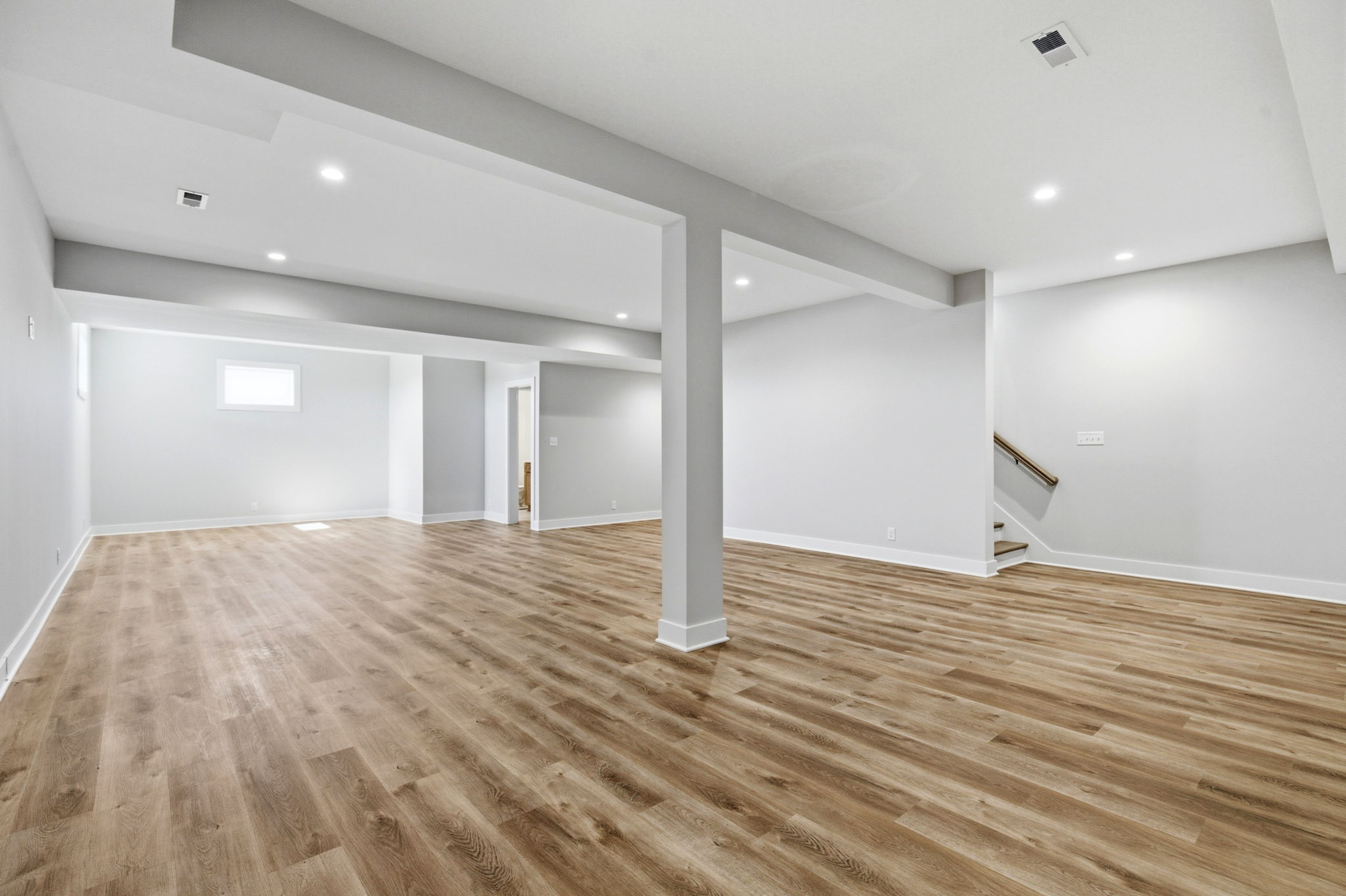 Open, empty basement with light gray walls, wood-look flooring, exposed columns, and recessed ceiling lights.