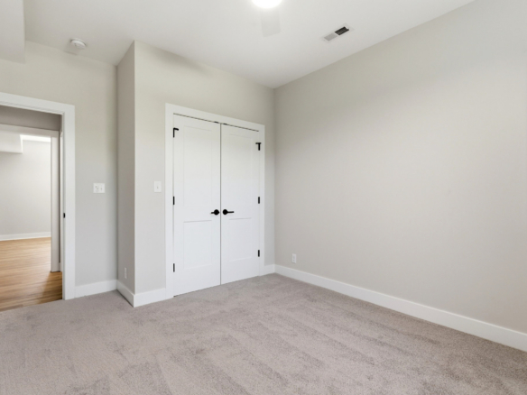 Empty neutral-toned bedroom with beige walls, white double closet doors, and light carpet. A doorway to a wood-floored room is visible.