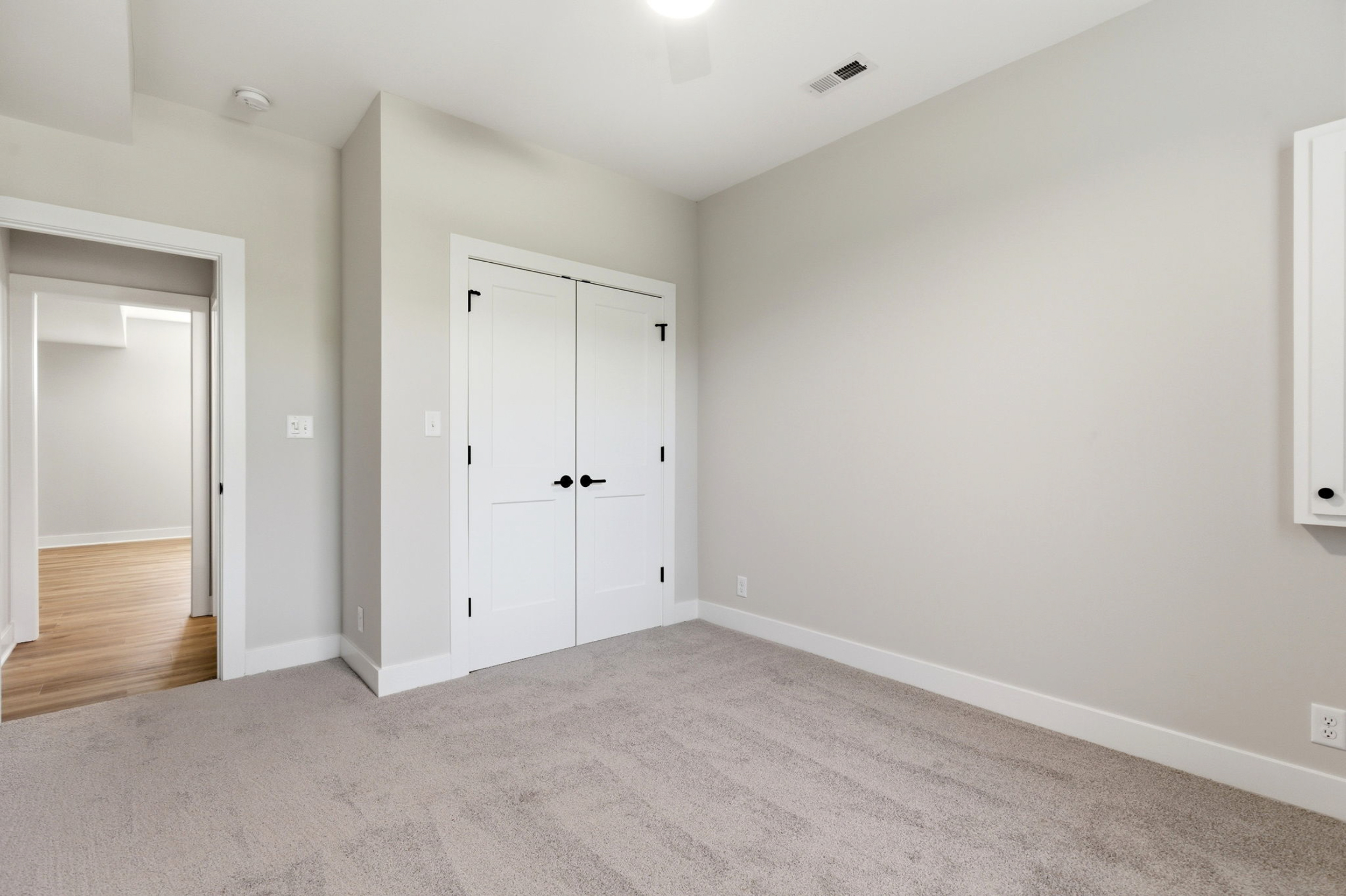 Empty neutral-toned bedroom with beige walls, white double closet doors, and light carpet. A doorway to a wood-floored room is visible.