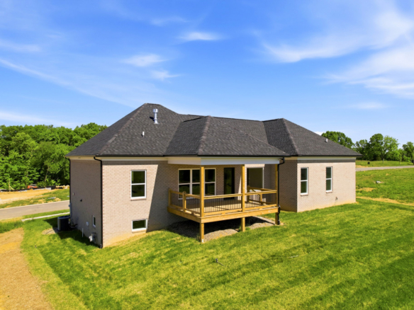 Single-story brick house with a wooden balcony, dark shingle roof, and a green lawn under a clear blue sky with trees in the background.