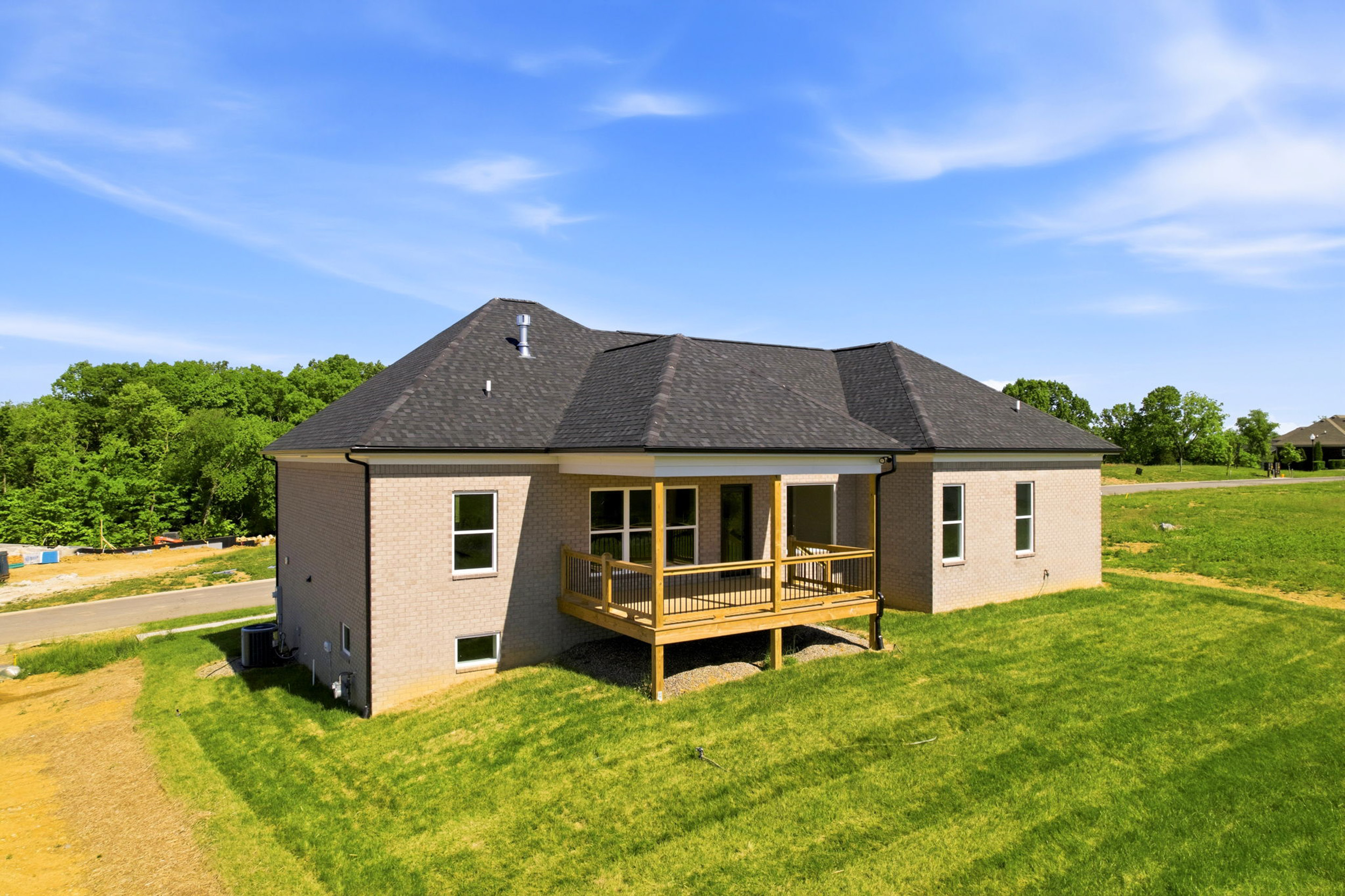 Single-story brick house with a wooden balcony, dark shingle roof, and a green lawn under a clear blue sky with trees in the background.