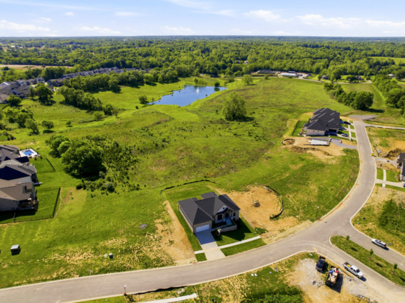 Aerial view of a developing suburban neighborhood with new houses along curved streets, green lawns, and a small central pond.