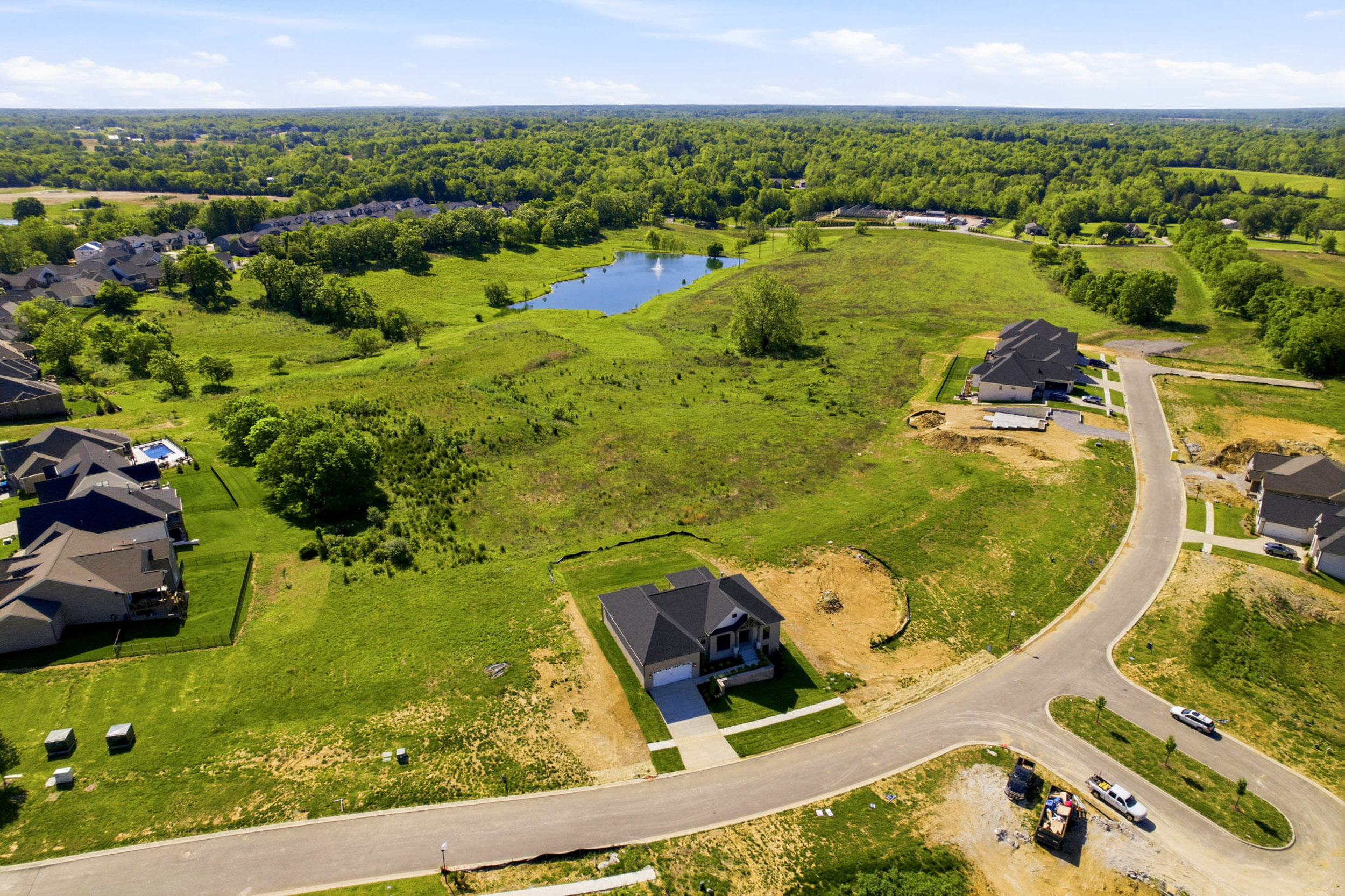 Aerial view of a developing suburban neighborhood with new houses along curved streets, green lawns, and a small central pond.
