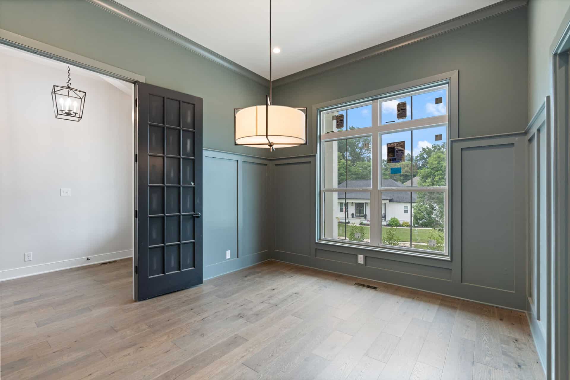 Room interior with a black paneled door open to a light-filled space and a large window showing a suburban view; muted green walls and a cylindrical hanging light fixture.