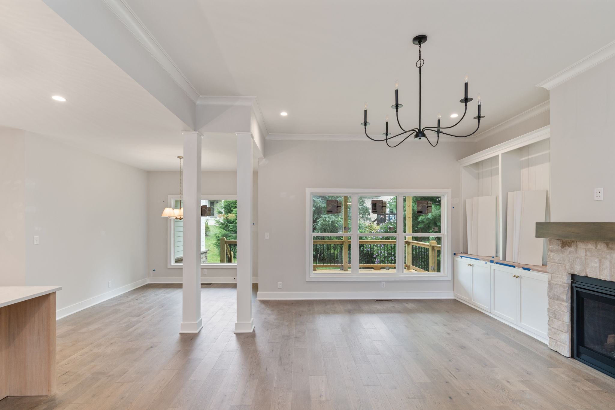 Open-concept living space with gray walls, white trim, a black multi-arm chandelier, large windows, and a fireplace with white cabinets beside.