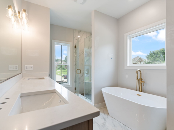 Bright modern bathroom with a white marble countertop, dual sinks, glass shower, and freestanding tub by a large window.