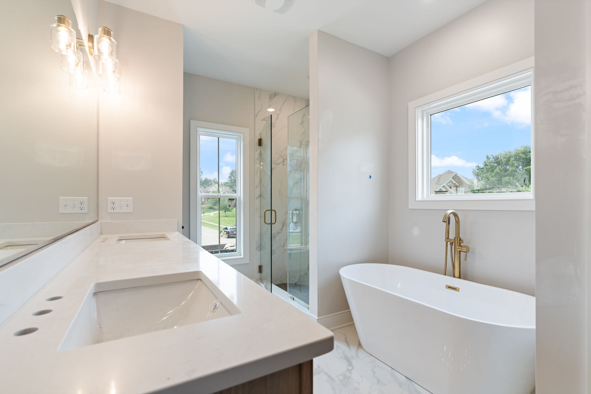 Bright modern bathroom with a white marble countertop, dual sinks, glass shower, and freestanding tub by a large window.