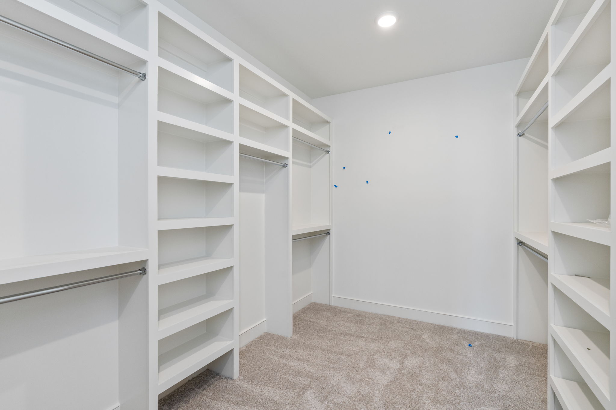 Walk-in closet with white built-in shelving units and metal clothing rods, beige carpet beneath and bright overhead light.