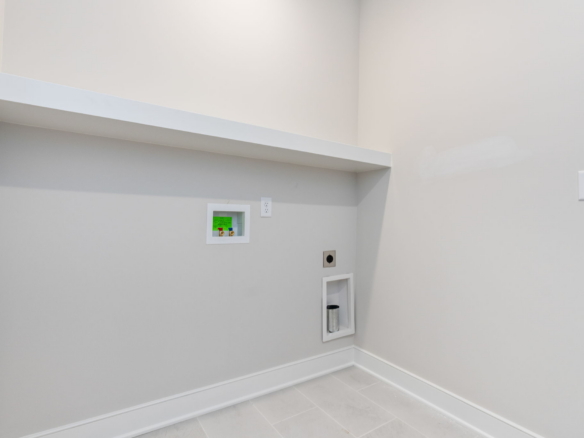 Utility room with light gray walls, a white diagonal shelf, and recessed electrical outlets and panels along the wall. A tall metal vent/pipe extends into a small recessed cavity near the floor. Clean, empty space with tile floor.