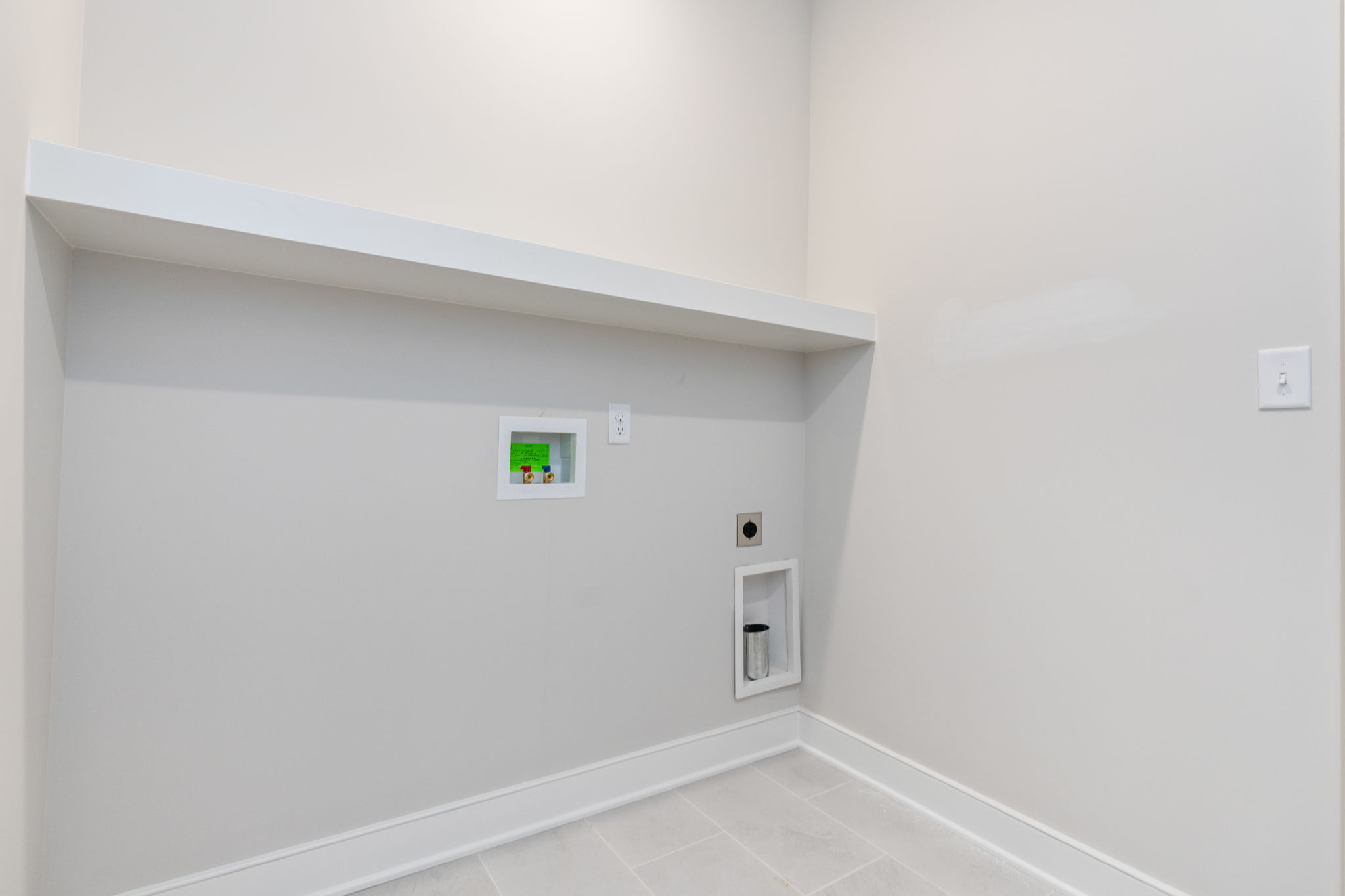 Utility room with light gray walls, a white diagonal shelf, and recessed electrical outlets and panels along the wall. A tall metal vent/pipe extends into a small recessed cavity near the floor. Clean, empty space with tile floor.