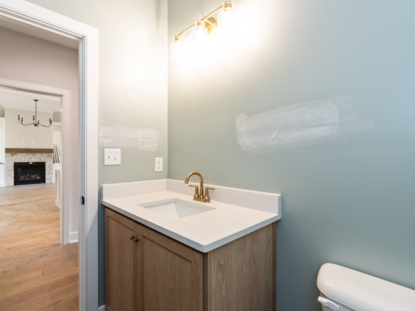 Bathroom vanity with white countertop and brass faucet against pale blue walls; doorway leads to living room with fireplace in background.