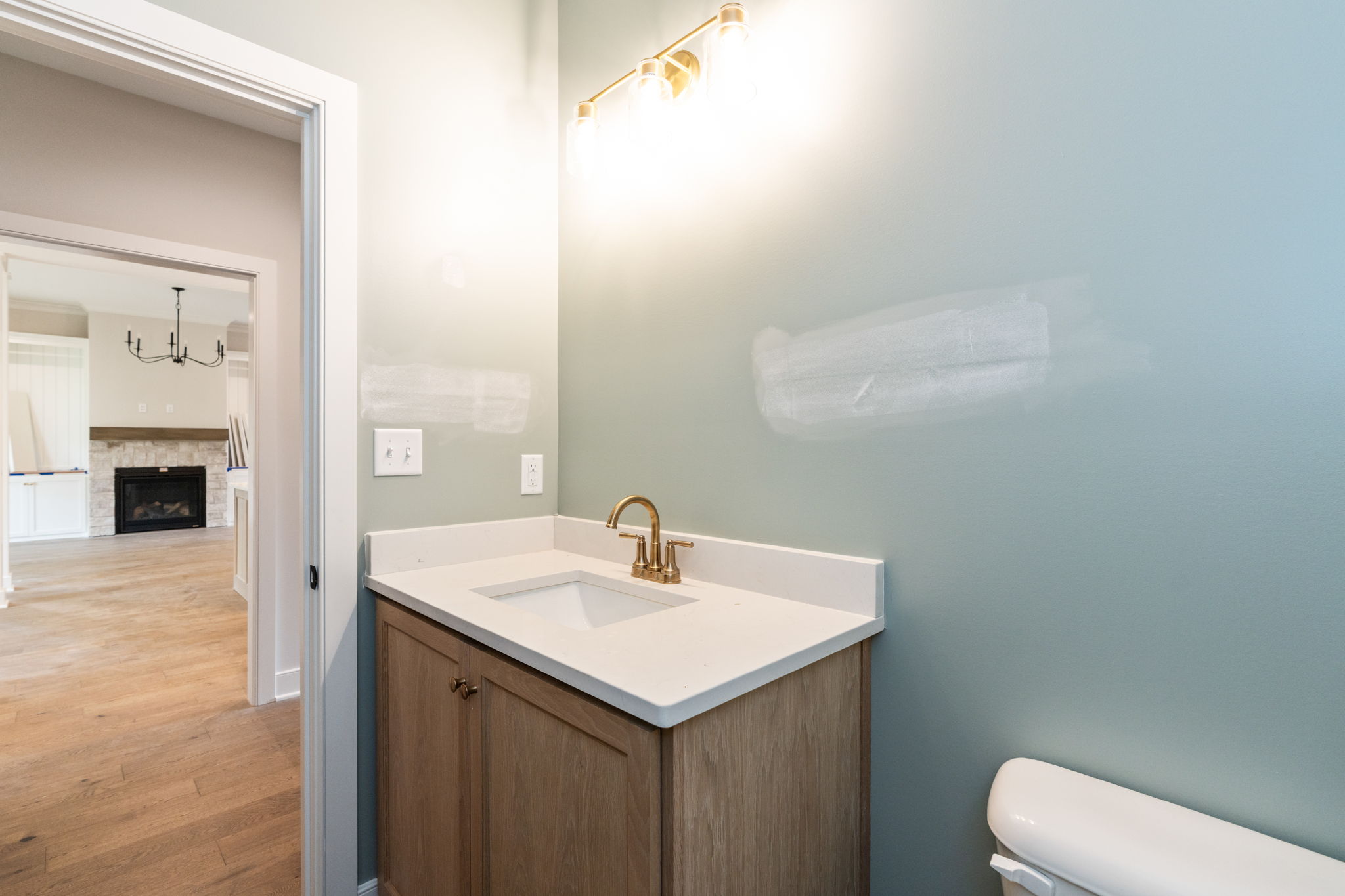 Bathroom vanity with white countertop and brass faucet against pale blue walls; doorway leads to living room with fireplace in background.