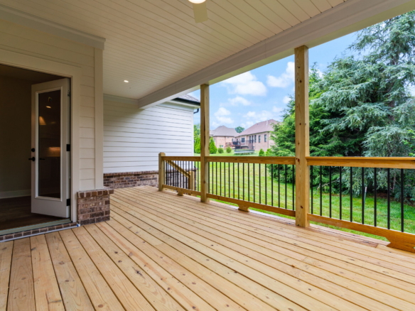 Open back porch with light-colored siding, wooden decking, and a railing overlooking a green yard and neighboring houses.
