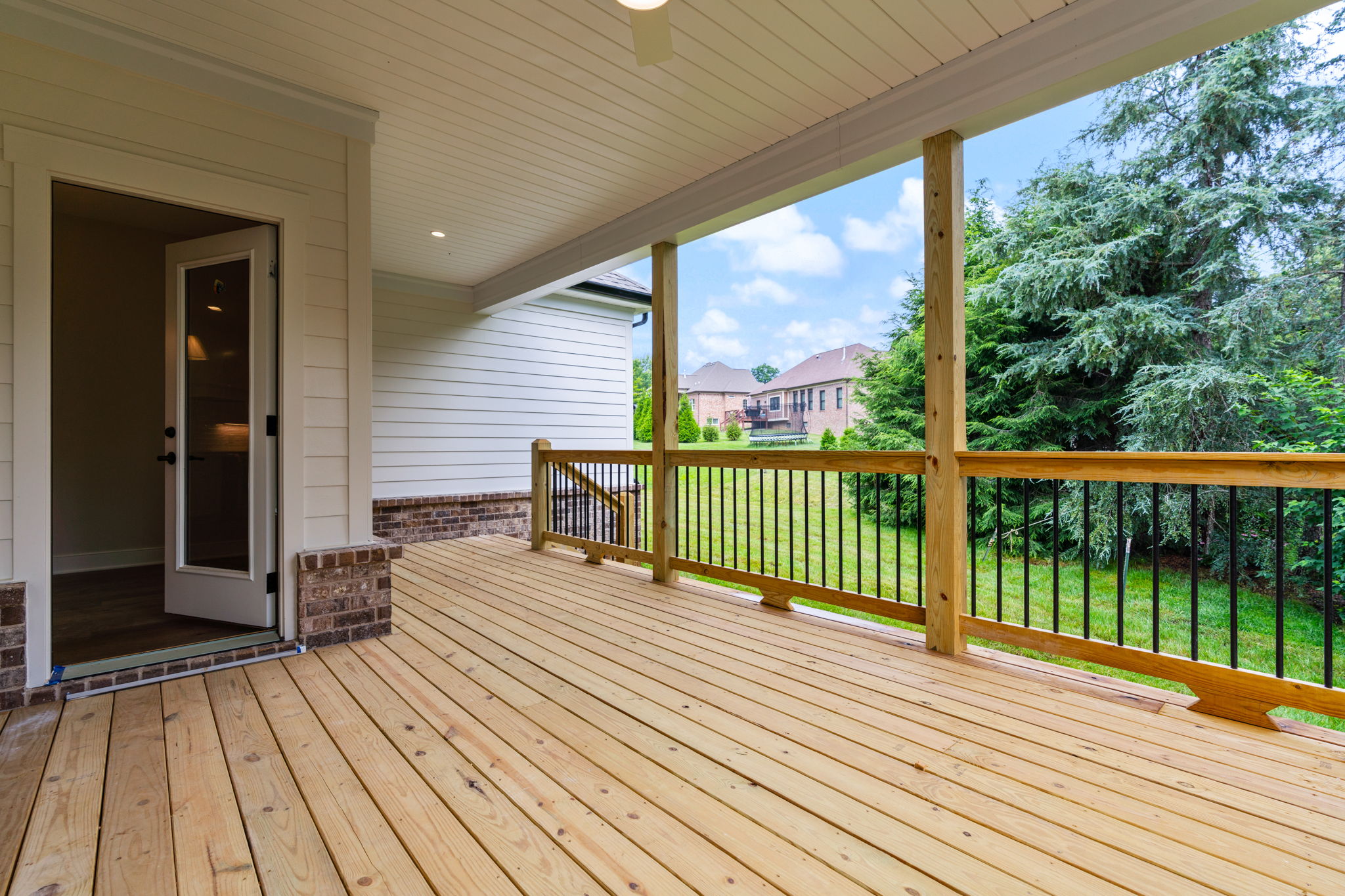 Open back porch with light-colored siding, wooden decking, and a railing overlooking a green yard and neighboring houses.