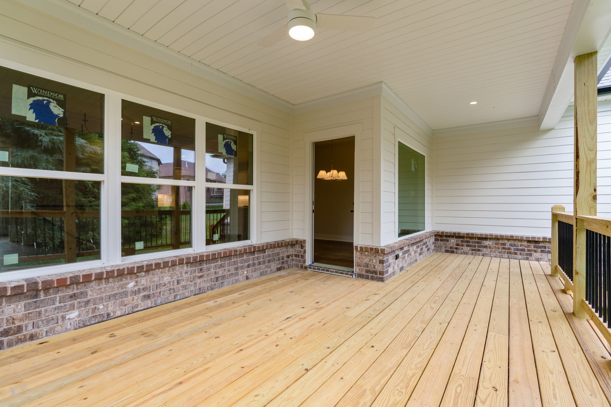 Covered wooden porch with brick base, large windows, and an open door revealing a chandelier inside.