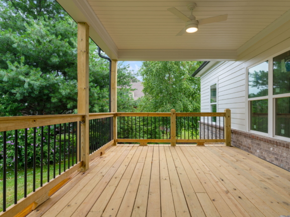 Wooden deck with black railing beside a white house, surrounded by lush green trees in the yard.