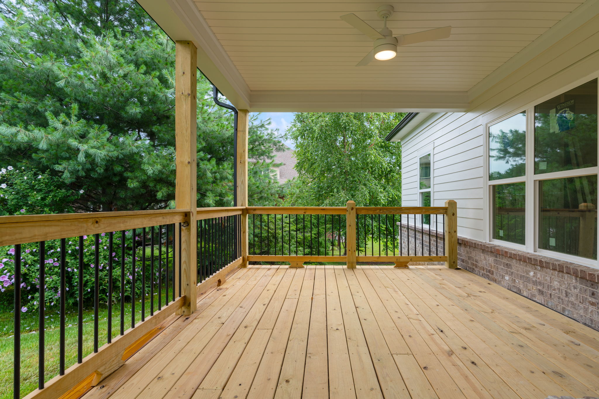 Wooden deck with black railing beside a white house, surrounded by lush green trees in the yard.