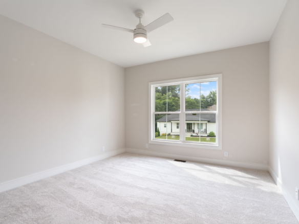 Empty bright bedroom with beige walls, light carpet, and a white ceiling fan; window shows a neighborhood view.