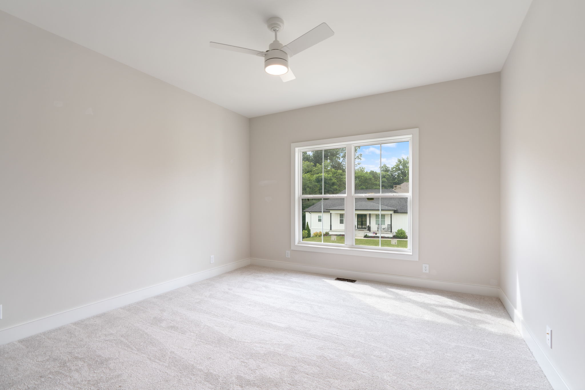 Empty bright bedroom with beige walls, light carpet, and a white ceiling fan; window shows a neighborhood view.