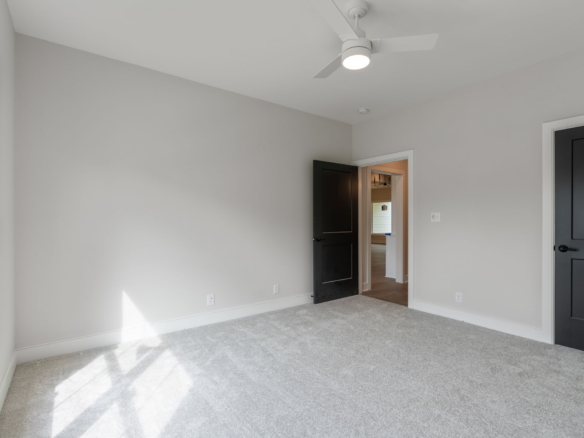 Empty bedroom with light gray walls, white ceiling fan, and light beige carpet; open door shows adjoining room.