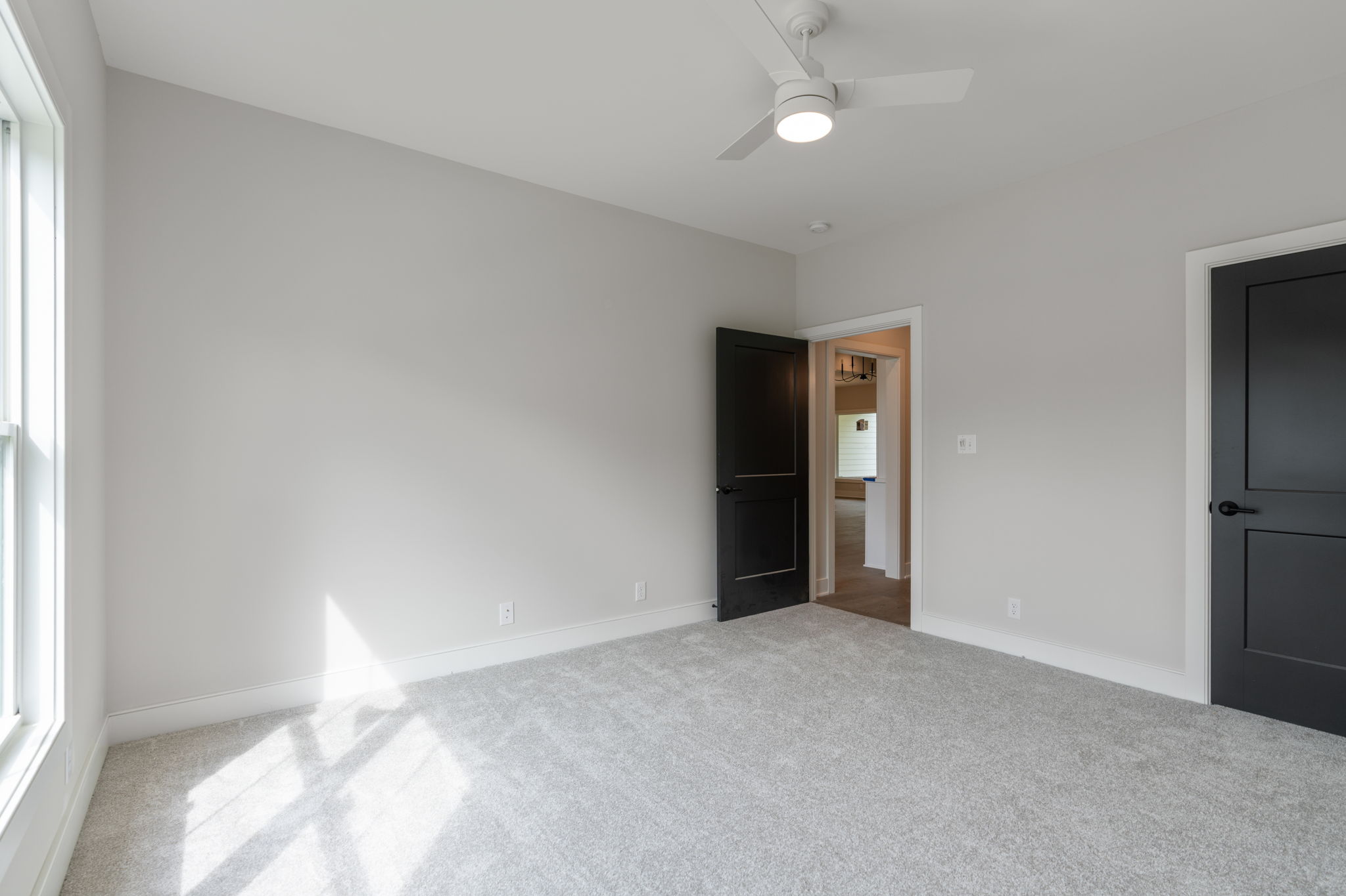 Empty bedroom with light gray walls, white ceiling fan, and light beige carpet; open door shows adjoining room.