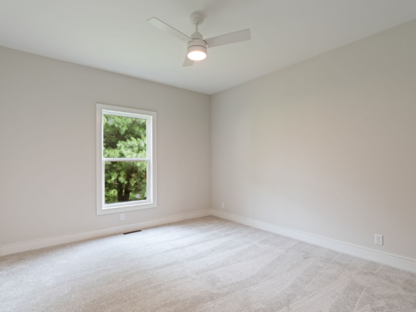 Empty bedroom with beige walls, white window trim, carpeted floor, and a white ceiling fan with light in the center