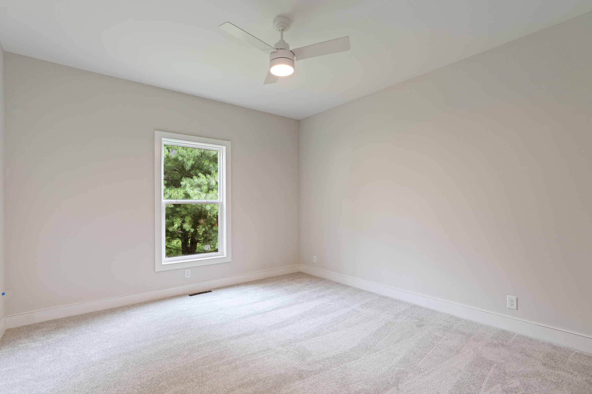 Empty bedroom with beige walls, white window trim, carpeted floor, and a white ceiling fan with light in the center