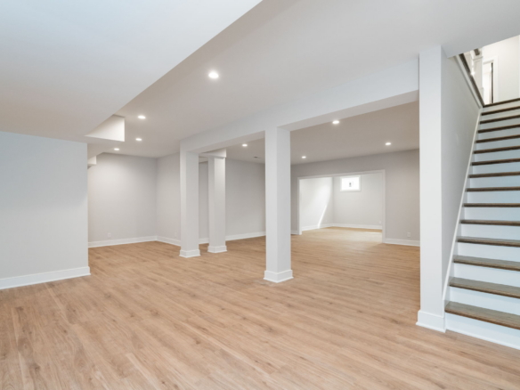 Finished basement with light wood flooring, white walls, recessed lights, and a wooden staircase along the right side.