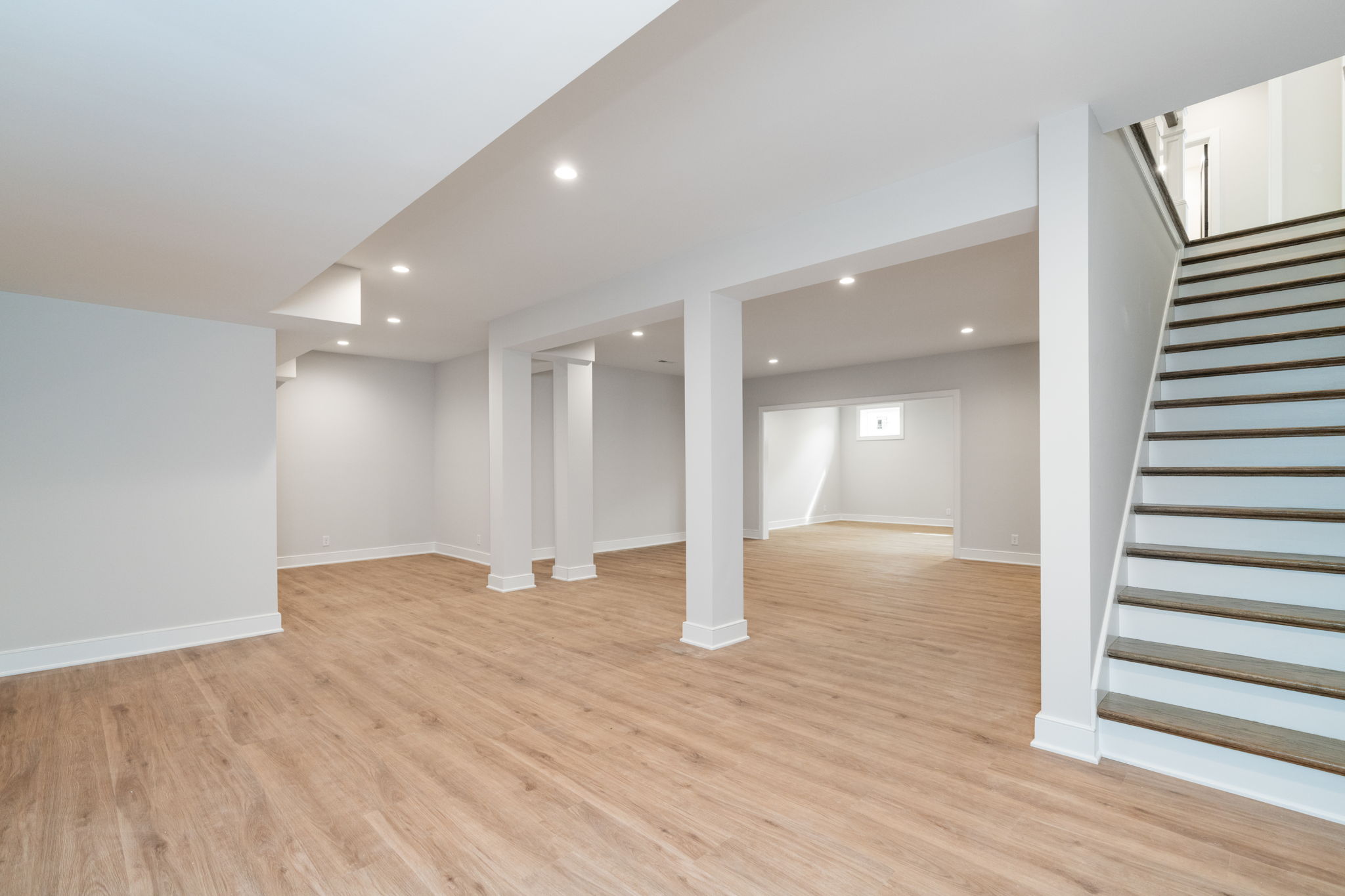 Finished basement with light wood flooring, white walls, recessed lights, and a wooden staircase along the right side.