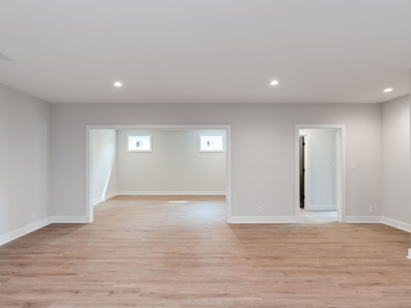 Empty, unfinished-style living space with light gray walls, wood-look flooring, and recessed ceiling lights; doorways into adjacent rooms visible at the far wall.