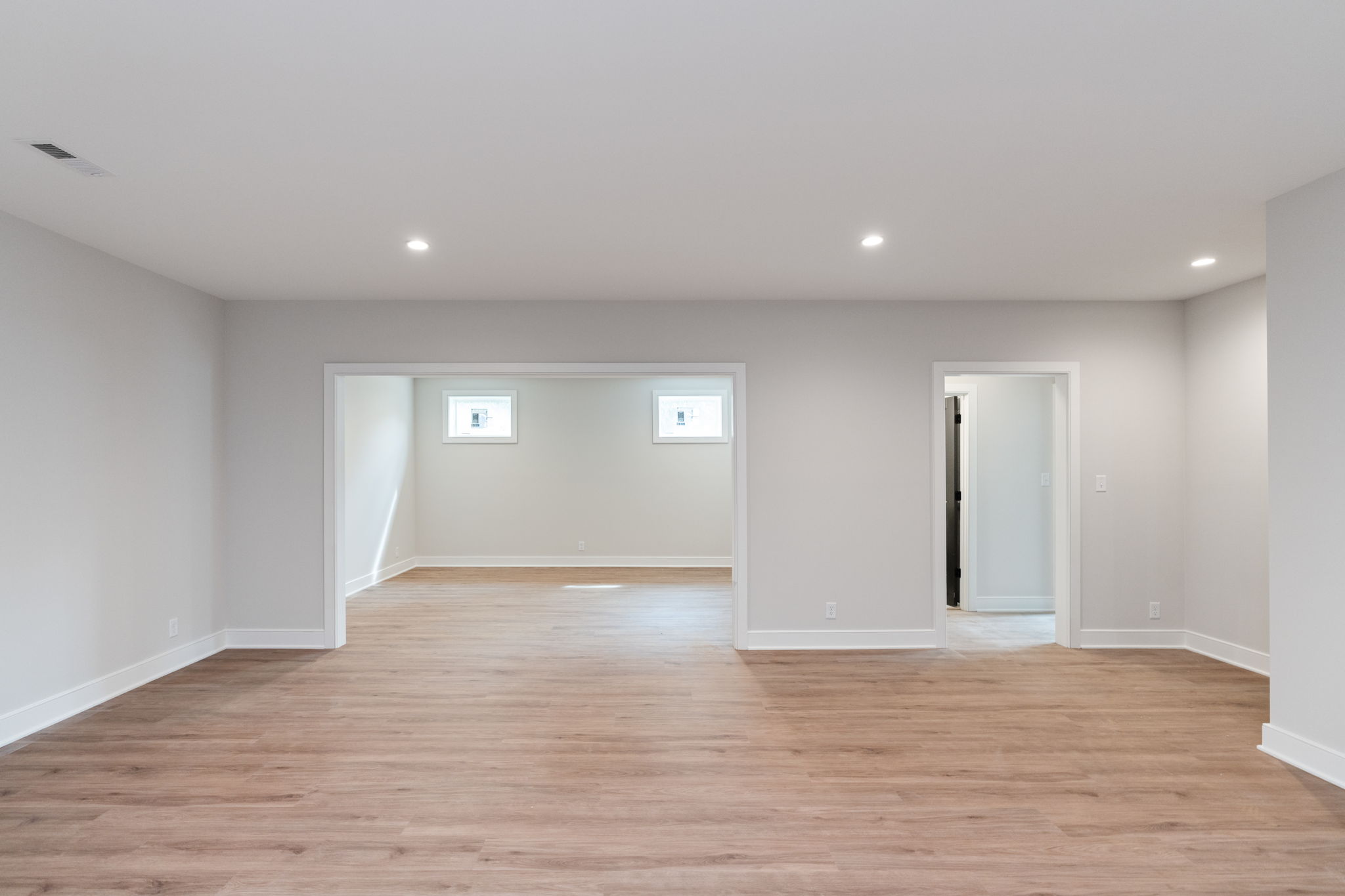 Empty, unfinished-style living space with light gray walls, wood-look flooring, and recessed ceiling lights; doorways into adjacent rooms visible at the far wall.