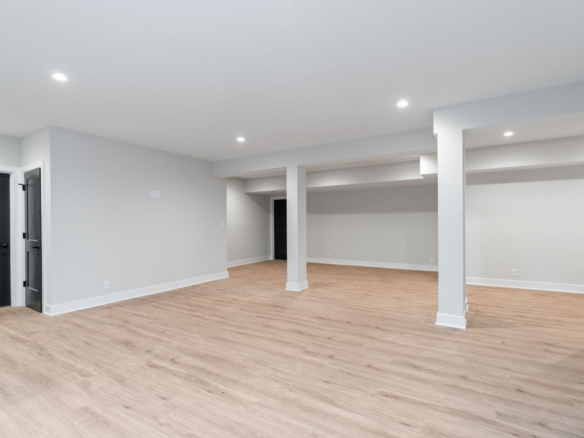 Empty finished basement with light gray walls, white trims, wood-look flooring, and multiple support columns.