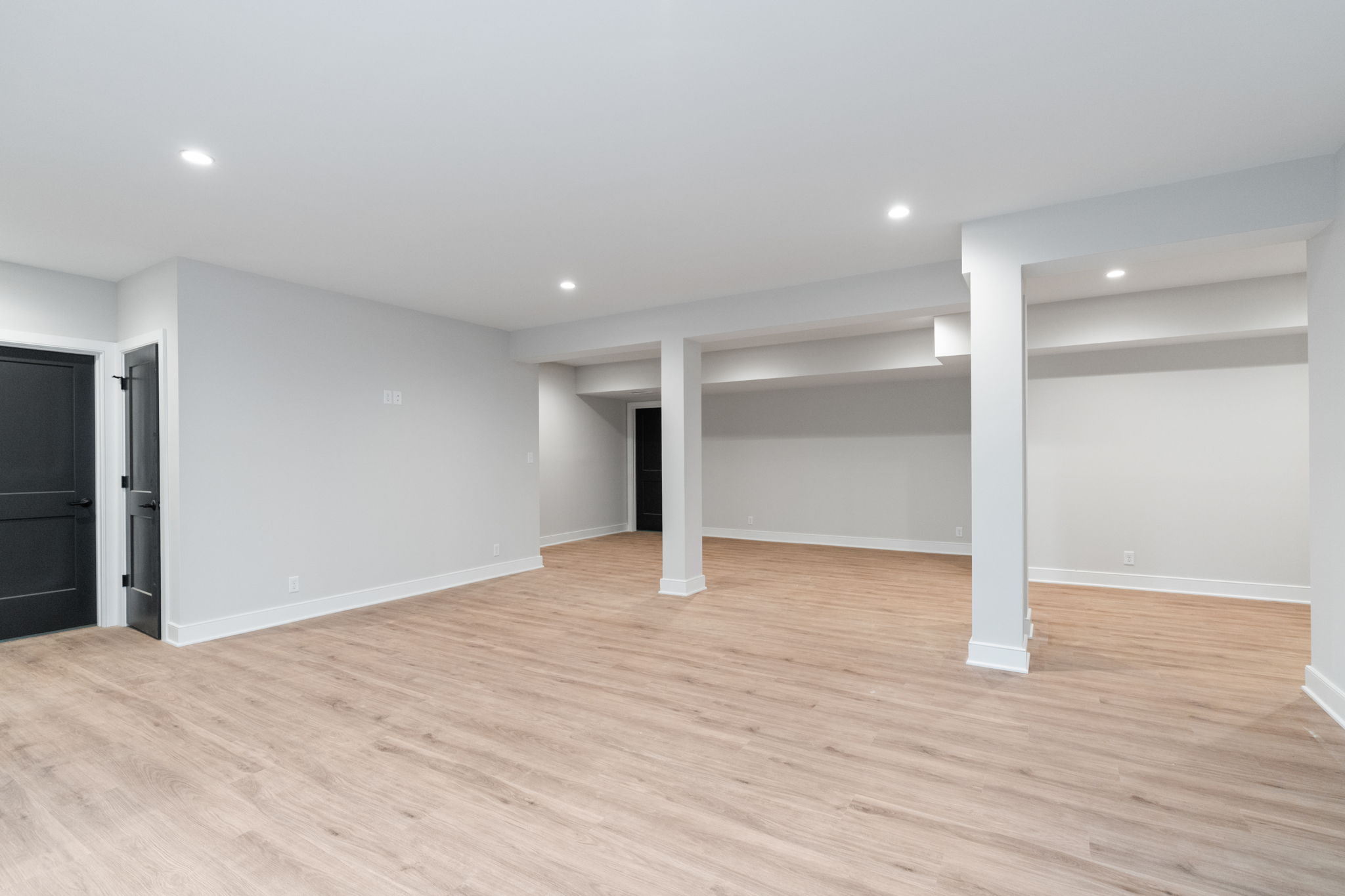Empty finished basement with light gray walls, white trims, wood-look flooring, and multiple support columns.