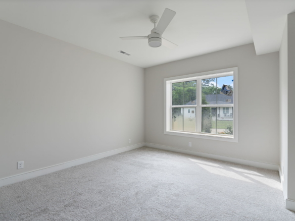 Empty light-gray bedroom with a large window and ceiling fan, carpeted floor, and neutral walls awaiting furnishings.