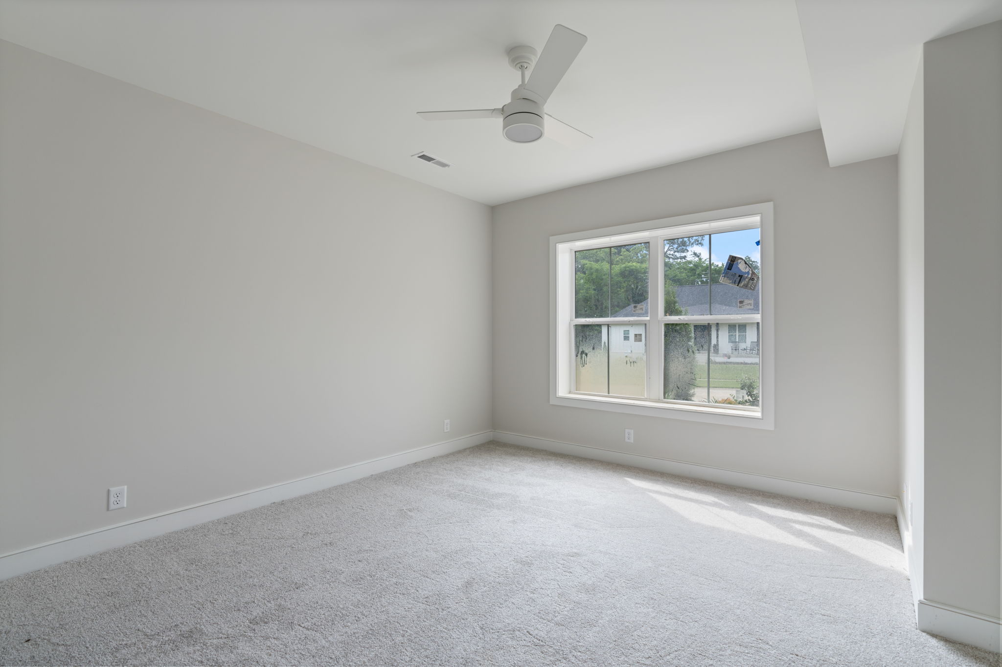 Empty light-gray bedroom with a large window and ceiling fan, carpeted floor, and neutral walls awaiting furnishings.