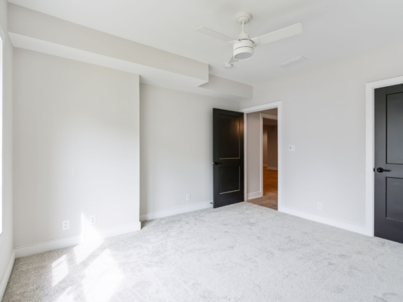 Empty modern bedroom with light gray walls, white trim, and a white ceiling fan mounted to the ceiling, with carpeted floor and an open dark door to another room.