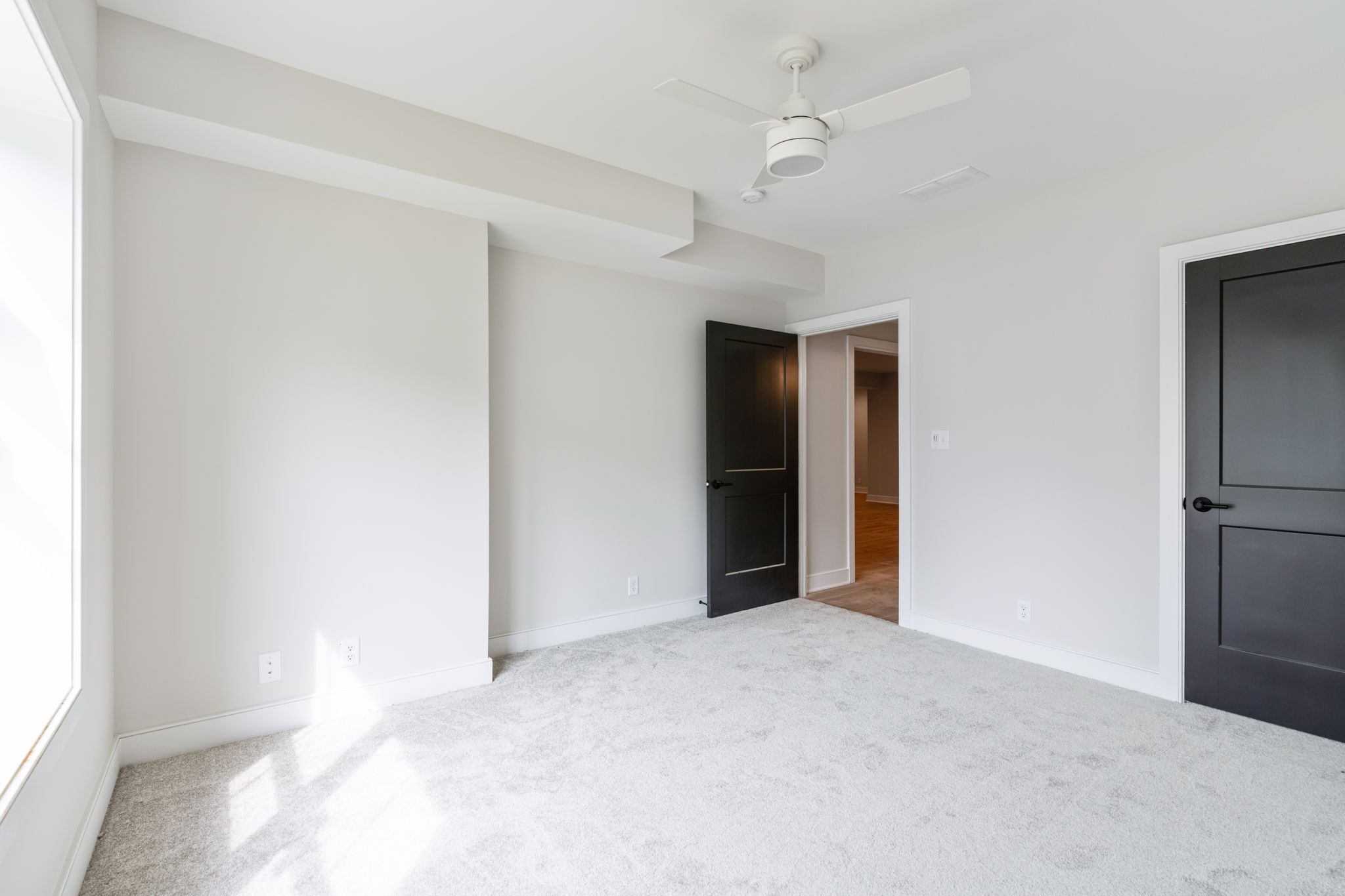Empty modern bedroom with light gray walls, white trim, and a white ceiling fan mounted to the ceiling, with carpeted floor and an open dark door to another room.