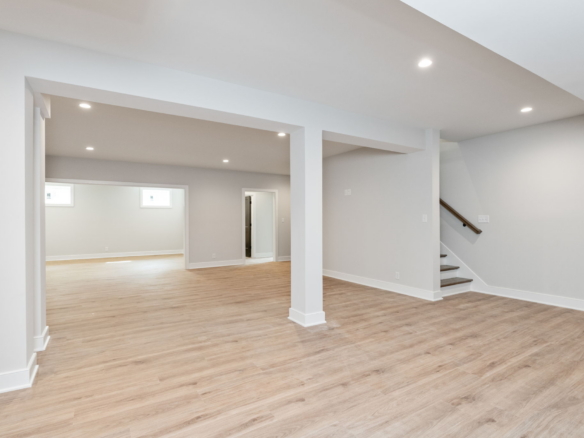 Open basement living area with white columns, light gray walls, and light wood flooring; staircase to the right.