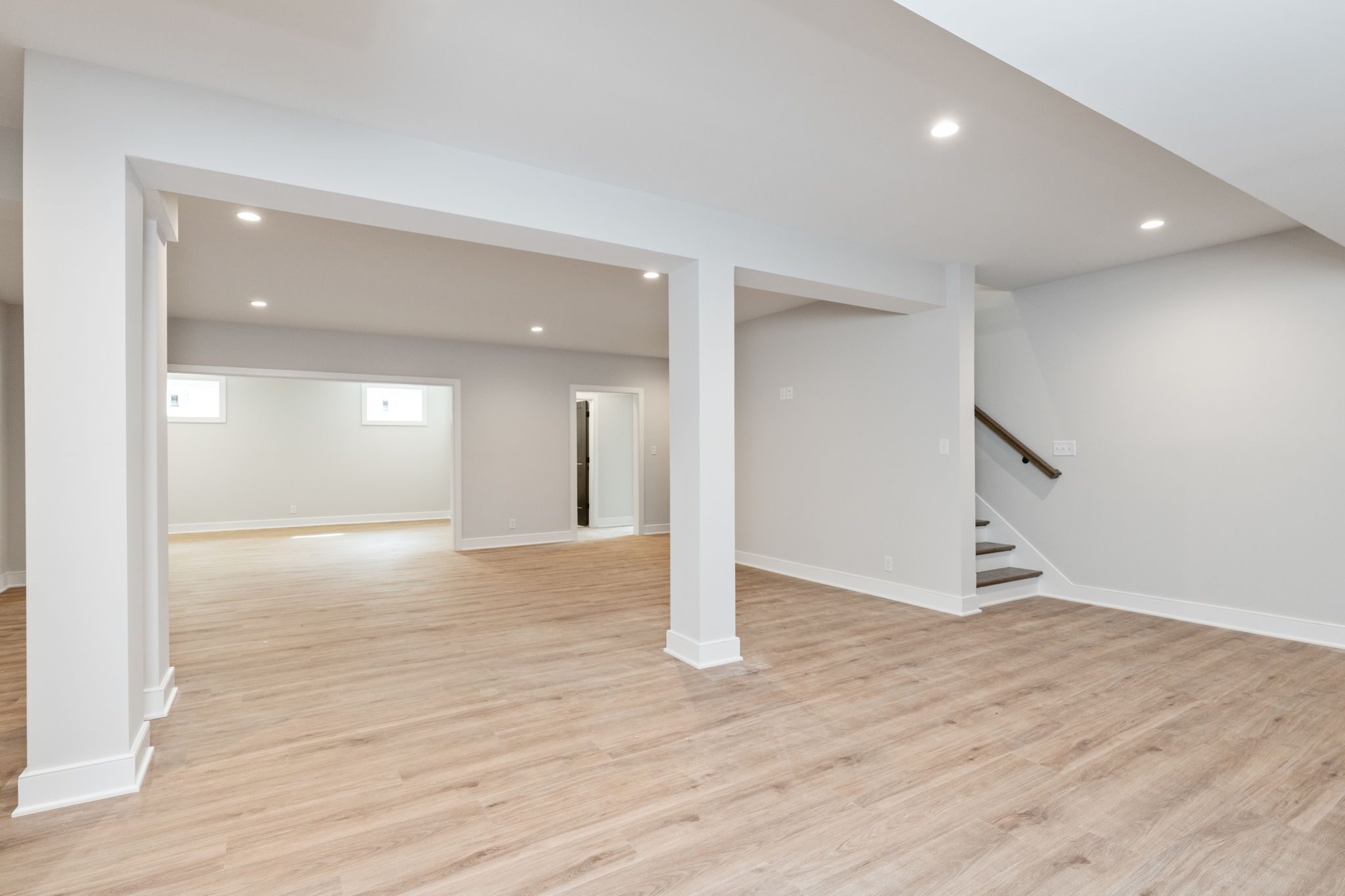 Open basement living area with white columns, light gray walls, and light wood flooring; staircase to the right.
