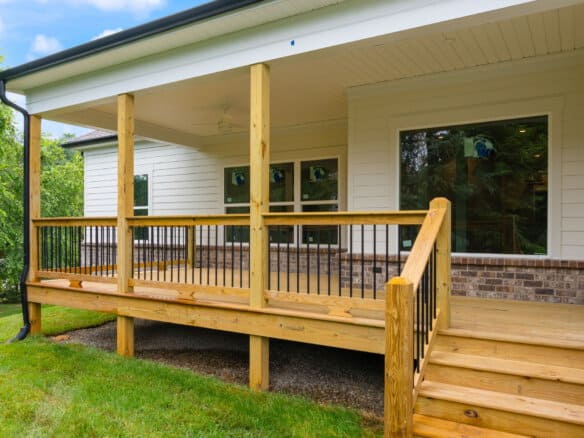 Front porch of a light-colored house with wooden railing, steps, and large windows reflecting green trees in the yard.